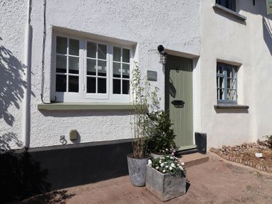Exterior of a white stucco cottage with green door and two windows with flower pots outside at Robyn Cottage Otterton