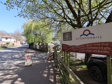 Roadside signs for Otterton Mill cafe restaurant and shops near a tree and parked car in Otterton