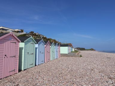 A row of pastel-colored beach huts on a pebble beach with houses on a hill in the background at Robyn Cottage in Otterton