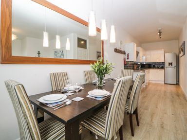 A dining area with a dark wooden table and six striped chairs set with plates and glasses next to a kitchen with white cabinets and appliances at Quayside Lookout in Brixham