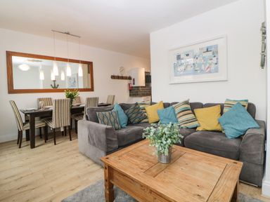 A living room with a grey sectional sofa with colorful cushions a wooden coffee table with a plant and a dining area with a table and striped chairs at Quayside Lookout in Brixham