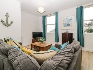 A living room with a gray sofa and colorful cushions a wooden coffee table a television on a wooden stand and teal curtains at Quayside Lookout in Brixham