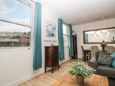 A living room with a gray sofa and wooden coffee table with a plant a dining table with striped chairs and a large mirror on the wall at Quayside Lookout in Brixham