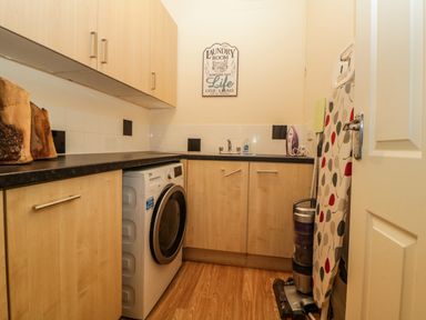 A laundry room with washing machine wooden cabinets ironing board and vacuum cleaner at Quayside Lookout in Brixham