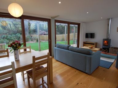 A living room with a wooden dining table and chairs a blue sofa a wood burning stove and large glass doors overlooking a garden at Orchard Retreat in Sidmouth