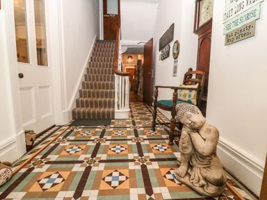 A hallway with patterned tiled floor a striped carpeted staircase a wooden chair with cushions a statue a grandfather clock and wall signs at Muriau Park in Caernarfon
