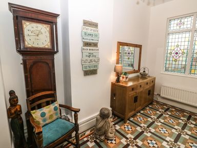 An entryway with a wooden chair, antique clock, wooden sideboard with lamp and mirror, stained glass window, and patterned floor tiles at Muriau Park in Caernarfon