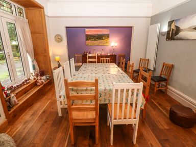 A dining room with a long table covered by a patterned tablecloth surrounded by mixed wooden chairs next to a window with stuffed animals on the ledge at Muriau Park in Caernarfon