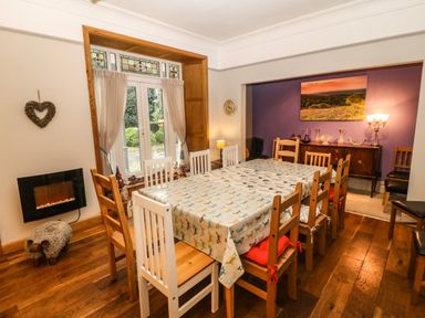 A dining room with a wooden table and chairs a window with curtains a sideboard with glassware and a wall with purple paint at Muriau Park in Caernarfon