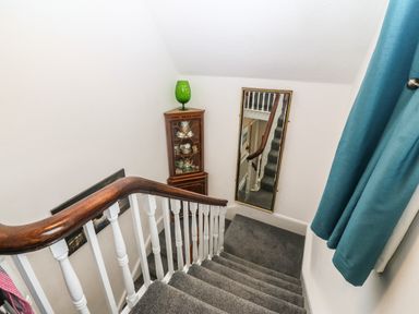 A carpeted staircase with a wooden handrail a tall mirror a corner cabinet with dishes and a green glass lamp and blue curtains at Muriau Park in Caernarfon