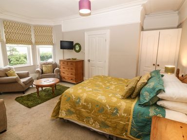 A bedroom with a bed and bedside table a chest of drawers a small round table two armchairs and windows with patterned blinds at Muriau Park in Caernarfon