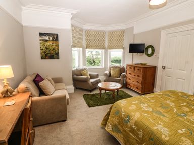 A bedroom with a bed a sofa two armchairs a round wooden table a wooden chest of drawers and a wooden desk at Muriau Park in Caernarfon