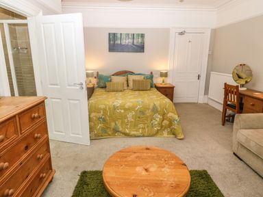 A bedroom with a bed with green and yellow bedding a wooden dresser a round wooden table a desk with a mirror and chair a door to a bathroom at Muriau Park in Caernarfon