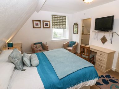 A bedroom with a bed covered in blue bedding two armchairs by a window a wooden desk and a wall-mounted television at Muriau Park in Caernarfon
