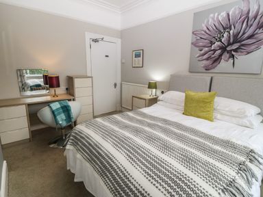 A bedroom with a bed a striped blanket a yellow pillow bedside tables lamps a desk chair a mirror a dresser and a large flower painting at Muriau Park in Caernarfon