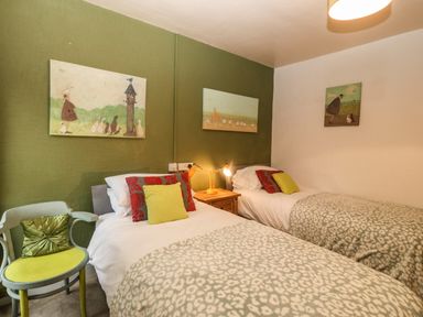 A bedroom with two single beds a wooden nightstand with lamps paintings on the walls a green chair with a cushion at Muriau Park in Caernarfon
