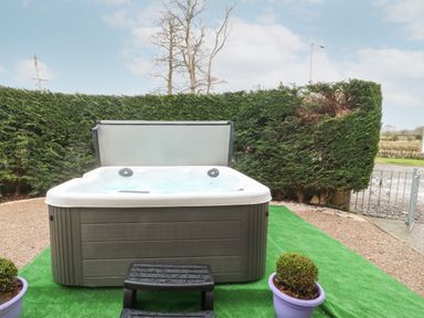 An outdoor hot tub with a cover partially open on a green artificial grass area with steps and two potted bushes at Muriau Park in Caernarfon