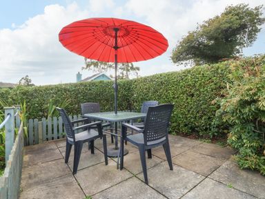 An outdoor patio with a table and four chairs under a red umbrella surrounded by hedges at 2 Argoed in Llanbedrog