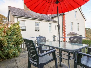 An outdoor patio with a glass table four black chairs and a large red umbrella near a white house at 2 Argoed in Llanbedrog