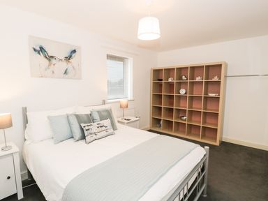 A bedroom with a bed and pillows a lamp on a bedside table a window and a large shelving unit at 7 Marina View in Pwllheli