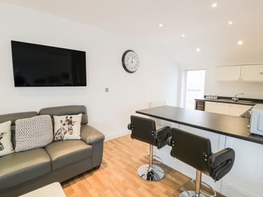 A living area with a gray sofa with animal print cushions a wall mounted TV a clock two black bar stools and a kitchen counter at 7 Marina View in Pwllheli