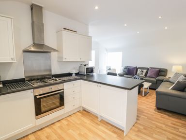 A kitchen with white cabinets black countertops and stainless steel oven next to a living area with gray sofas and a lamp at 7 Marina View in Pwllheli