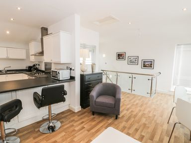 An open kitchen and seating area with black counter stools a gray armchair a black chest of drawers with a lamp and vase and framed pictures on a white wall at 7 Marina View in Pwllheli