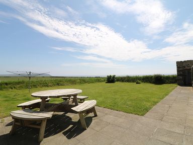 A patio area with a round wooden picnic table and benches next to a grassy lawn with a rotary clothesline and stone building at Trefollwyn Mawr in Abersoch