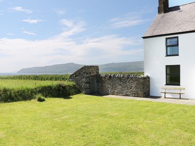 A grassy yard with a stone wall and a white house next to a bench overlooking hills and water at Trefollwyn Mawr in Abersoch