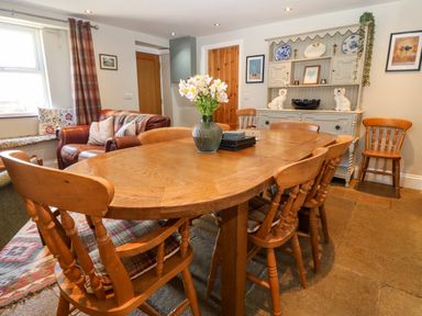 A dining room with wooden table and chairs a vase with flowers a cabinet with plates and decorative items and a sofa near a window at Trefollwyn Mawr in Abersoch
