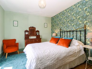 A bedroom with a metal bed frame white bedding orange and blue pillows wooden dresser with mirror orange armchair and patterned wallpaper at Trefollwyn Mawr in Abersoch