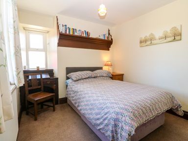A bedroom with a bed a wooden chair a small desk a bedside table with a lamp a bookshelf above the bed and a painting on the wall at Trefollwyn Mawr in Abersoch
