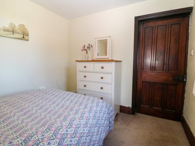 A bedroom with a bed covered in patterned bedding a white chest of drawers with a mirror and flowers and a wooden door at Trefollwyn Mawr in Abersoch