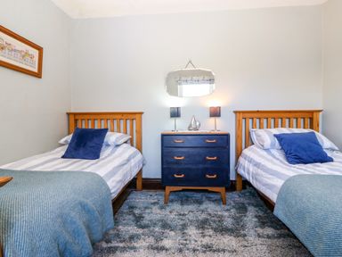 A bedroom with two single beds with striped bedding and blue pillows a blue chest of drawers with lamps and a mirror on the wall at Trefollwyn Mawr in Abersoch