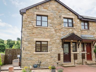 The exterior of a stone house with brown framed windows a dark brown door with a glass panel and a garden area with plants at 14 The Maltings in Rothbury