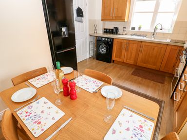 A kitchen with a wooden dining table set for four with plates glasses and a bottle next to wooden cabinets and a black refrigerator at 14 The Maltings in Rothbury