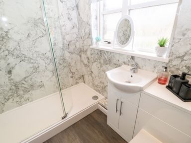A bathroom with a shower area with glass door white sink cabinets and window with plants and mirror at 14 The Maltings in Rothbury
