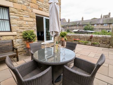 A patio with a round glass table and four wicker chairs an umbrella a wooden bench potted plants and a stone wall at 14 The Maltings in Rothbury