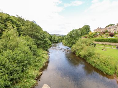 A river flowing between trees and bushes with houses and a grassy area on the right at 14 The Maltings in Rothbury