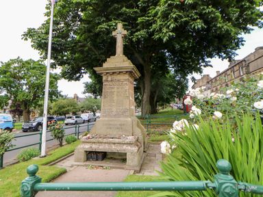 A war memorial monument with a cross at the top surrounded by flowers and greenery next to a street with parked cars and buildings in Rothbury