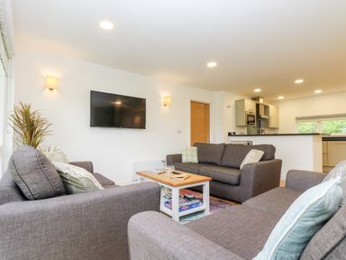 A living room with three gray sofas a wooden coffee table a wall mounted TV and a kitchen in the background at Beech in Truro