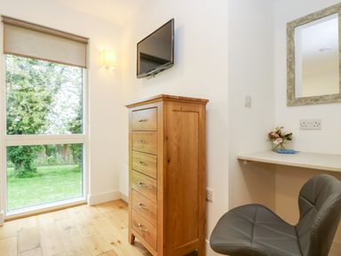 A corner of a room with a wooden chest of drawers a wall-mounted TV a window with a roller blind a countertop with flowers and a gray chair at Beech in Truro
