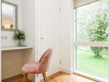 A room corner with a pink chair beside a white desk with flowers on it and a large window showing green trees at Beech in Truro