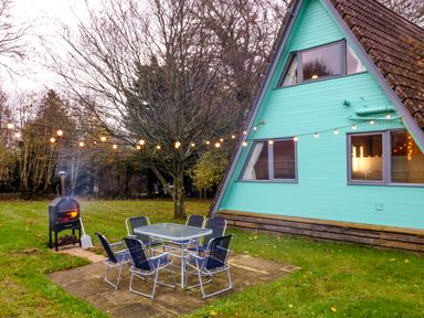 An outdoor patio with chairs and a table near a turquoise A-frame house with string lights and a wood stove