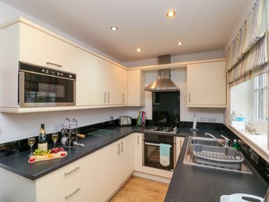 A kitchen with white cabinets black countertops a built-in oven a microwave a stainless steel sink and a window with striped blinds at Dotty's Sunshine Cottage The Bay near Filey