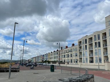 A promenade with parked cars and street lamps beside a row of multi-story buildings at Wavecrest in Pwllheli