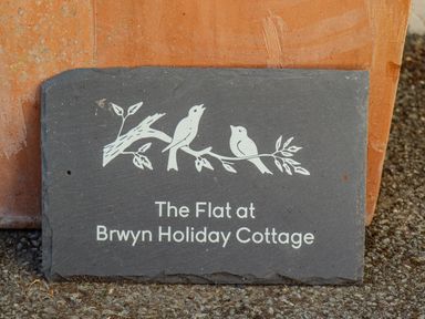 A slate sign with birds on a branch and text The Flat at Brwyn Holiday Cottage