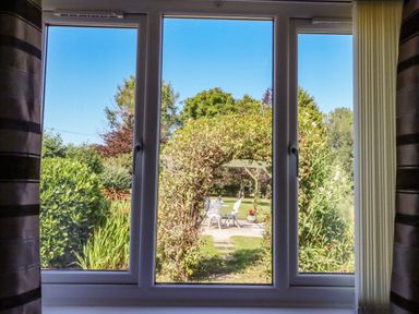 A window with curtains looking out to a garden with an archway and white chairs at The Flat At Brwyn in Brynford