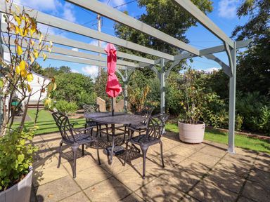 A patio with metal table and chairs under a pergola with a folded pink umbrella and plants in pots at The Flat At Brwyn in Brynford