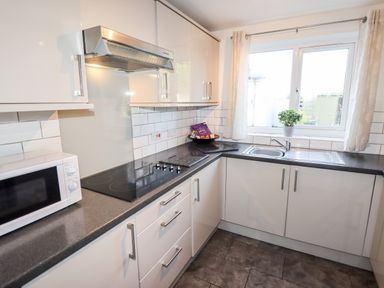 A kitchen with white cabinets a black countertop a microwave a stovetop a sink and a window with curtains and a plant at The Flat At Brwyn in Brynford